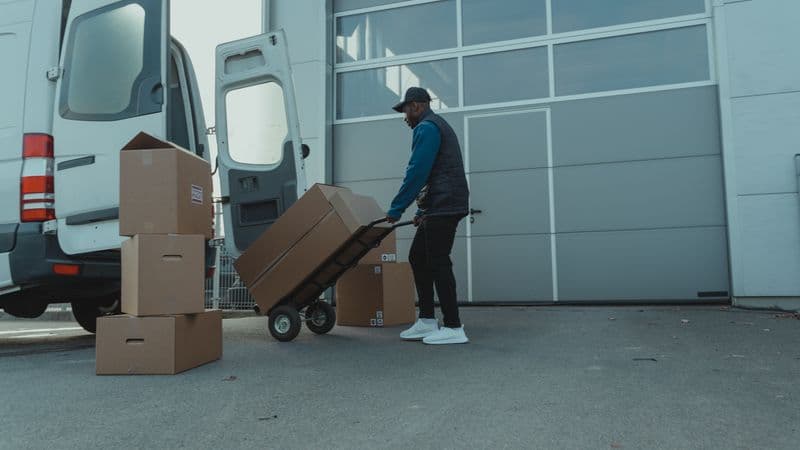 Kurir mendorong troli berisi kardus dari mobil box ke pintu gudang.